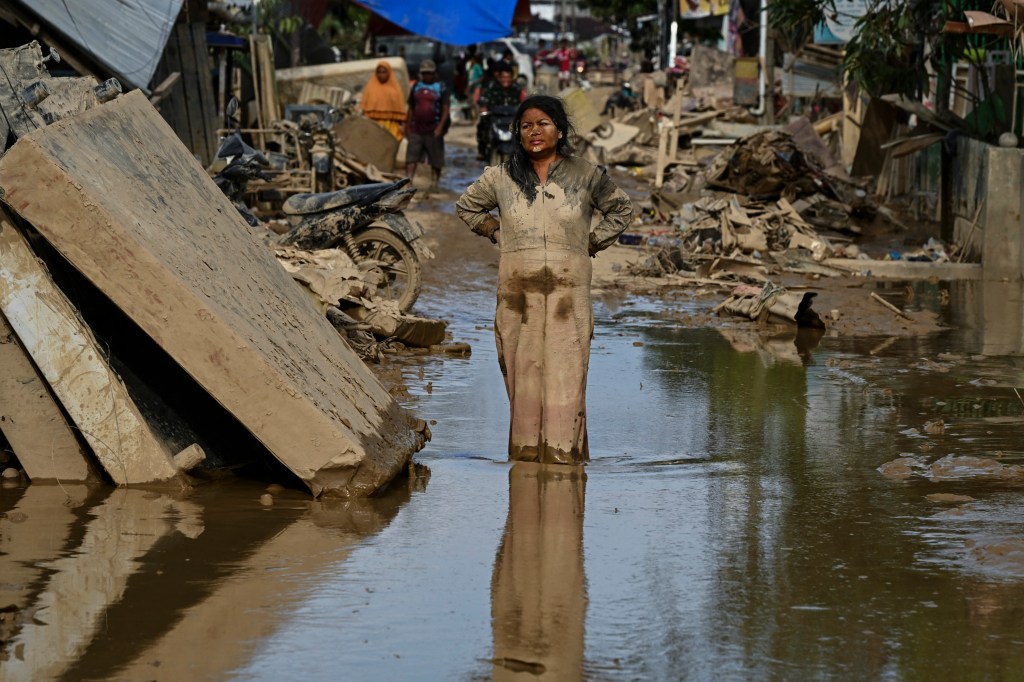 A woman covered in mud stands in a flooded, debris-filled street after a flash flood.