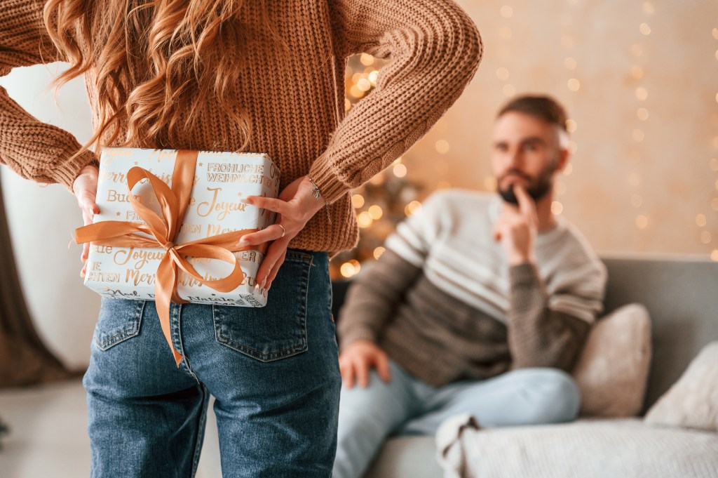 A woman hides a gift box behind her back for a man sitting on a couch.