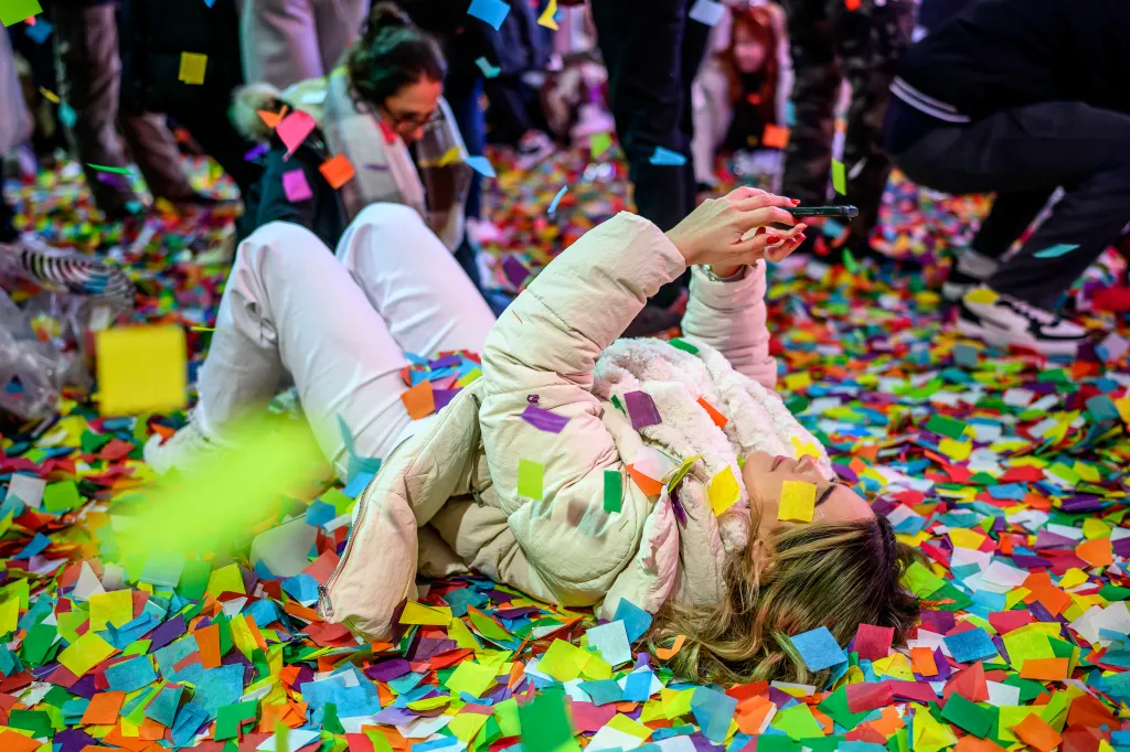Woman lying in colorful confetti, holding a smartphone up to take a photo or video.