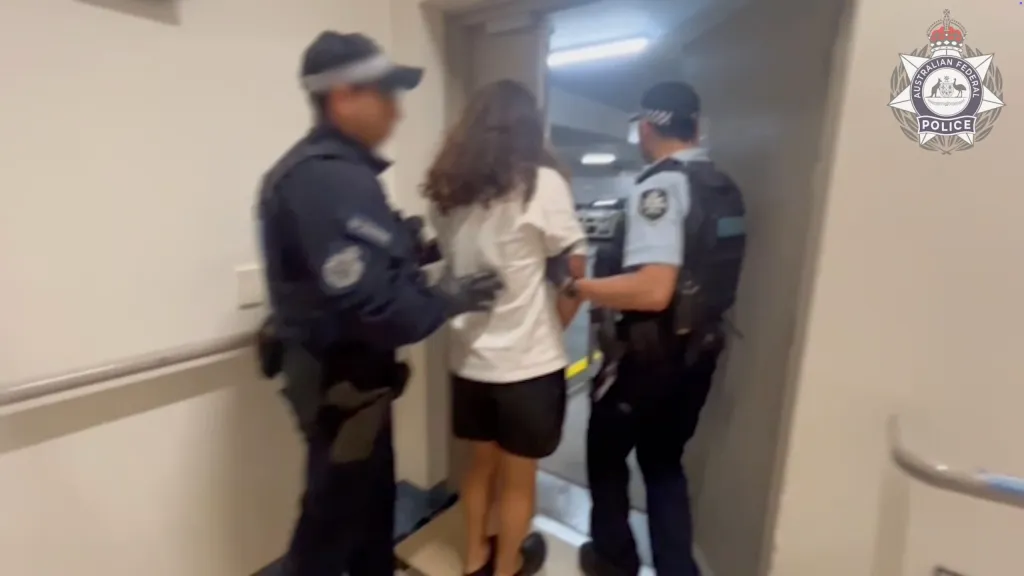 Two Australian Federal Police officers escorting a suspect wearing a white t-shirt and black shorts, with the AFP logo in the top right.