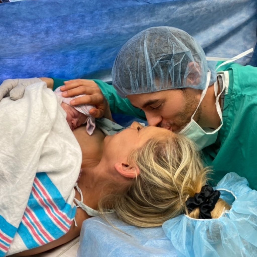 A man in medical scrubs and cap kisses a woman on the forehead as she holds a swaddled newborn.