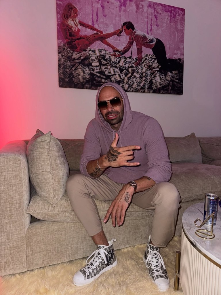 Gene Borrello sitting on a couch, holding up a hand sign with a painting of a man and woman on money in the background.
