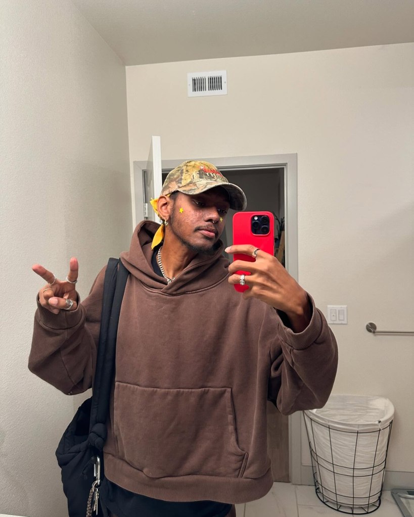 Niko McKnight wearing a brown hoodie, camouflage hat, and yellow bandana taking a mirror selfie.