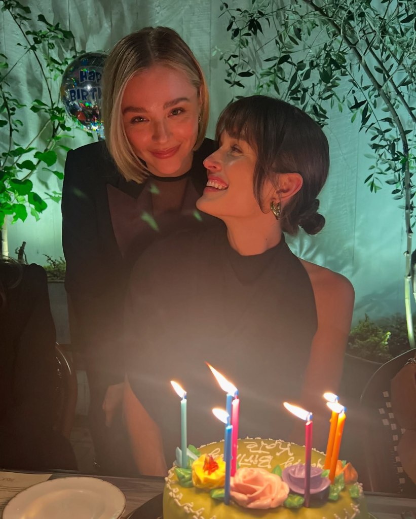 Chloe Grace Moretz looking at Kate Harrison, who is smiling and looking at her, with a birthday cake with lit candles in the foreground.