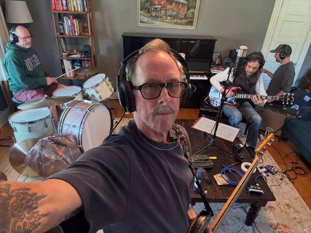 Weezer bassist Scott Shriner taking a selfie while playing bass with other musicians in a room.