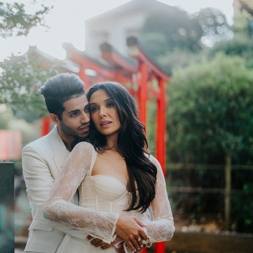 Mena Massoud embracing Emily Shah from behind in front of red torii gates.