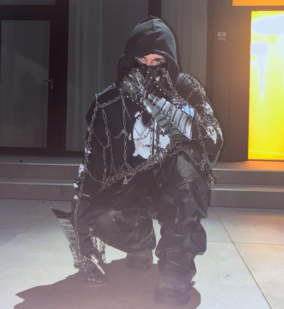 Person wearing a black hoodie, chains, armored gauntlets, and shiny black pants, crouching.