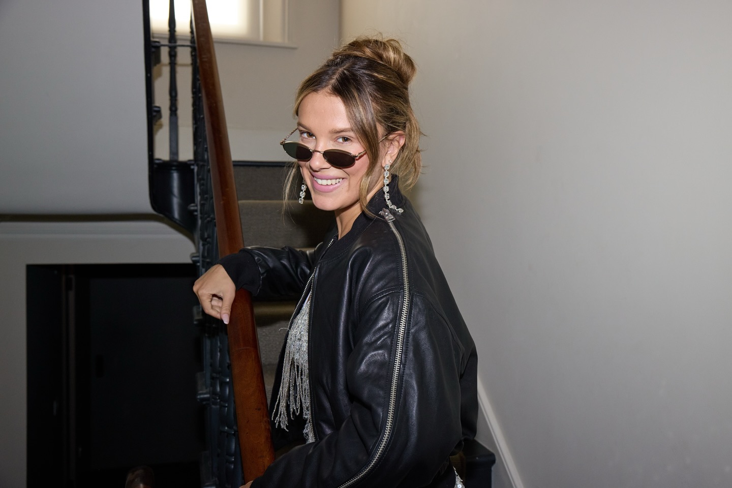 Millie Bobby Brown posing on a staircase wearing a black leather jacket, sunglasses, and large dangling earrings.