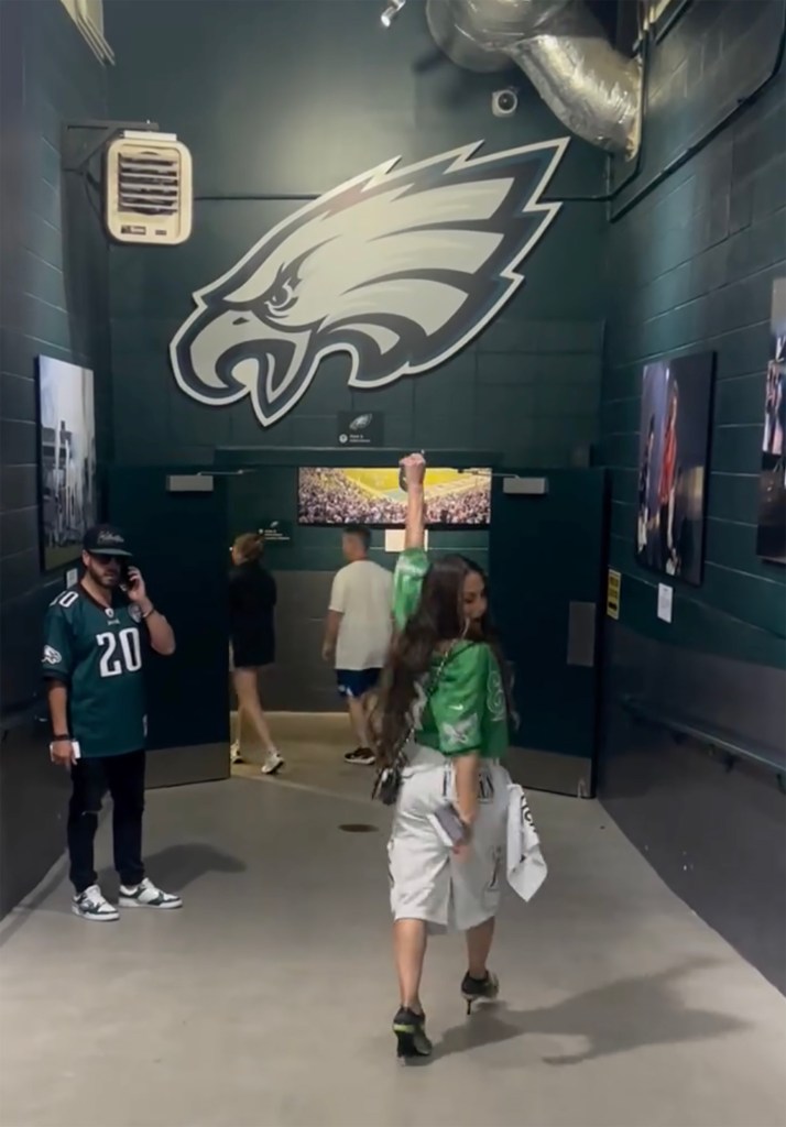 Nikki Bella, in a green Eagles jersey, walks through a stadium hallway with an Eagles logo on the wall.