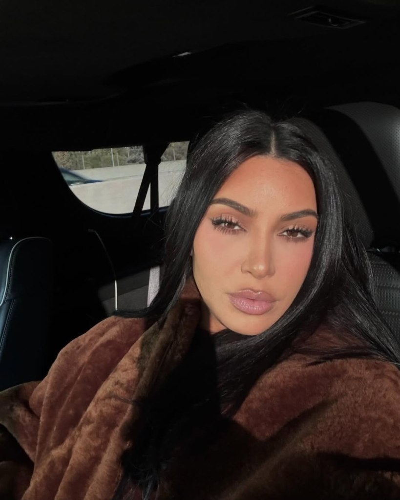 Kim Kardashian with long dark hair, wearing a brown furry coat, posing in a car.