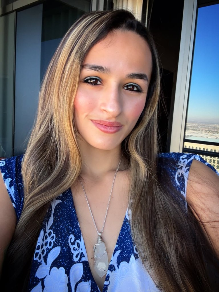 Jazz Jennings smiling, wearing a blue and white dress, with a necklace with a large crystal pendant.