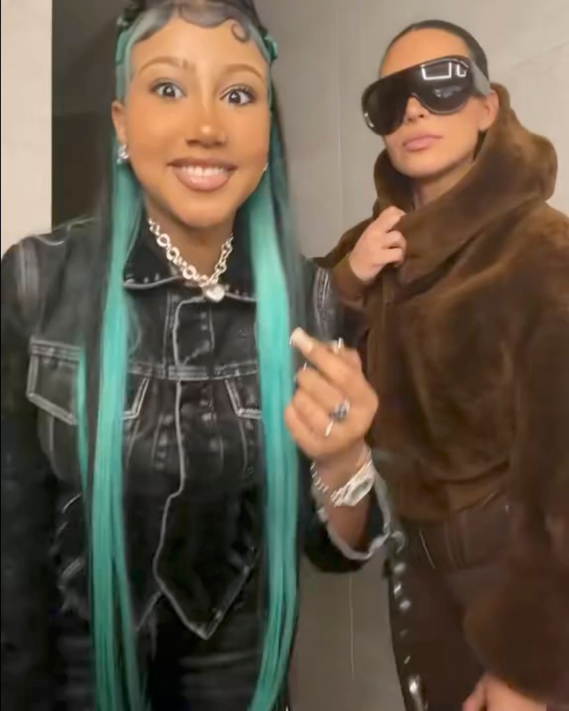 Two women, one with long turquoise hair making a silly face, and another with sunglasses wearing a brown furry hoodie, pose for a photo.