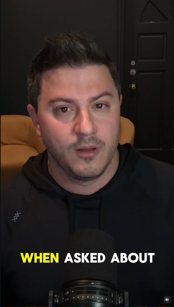 A man in a black hoodie speaks into a microphone.