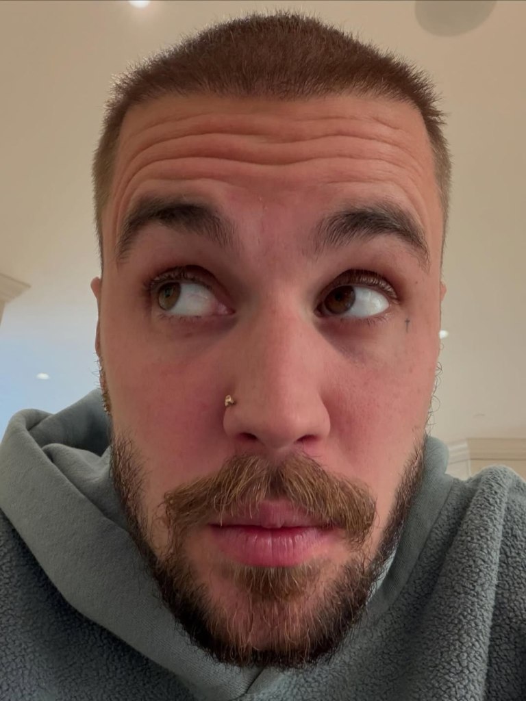 Justin Bieber wearing a grey hoodie and a nose ring, looking up.