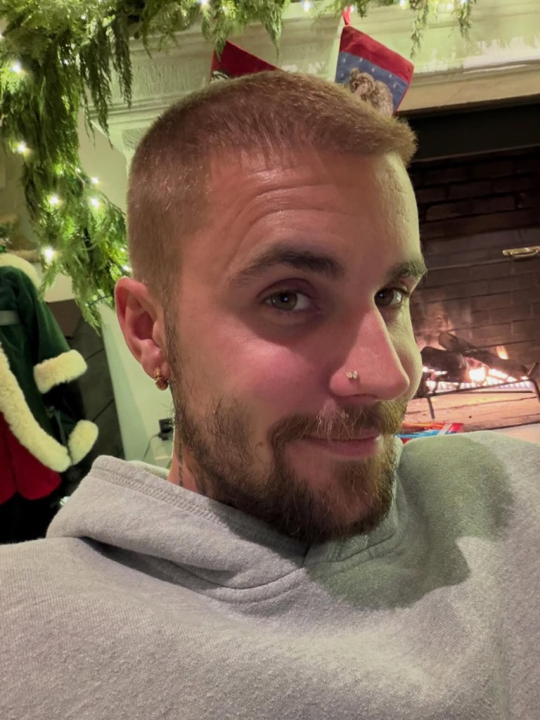 A man with a beard and short hair smiles, wearing a grey hoodie, with Christmas decorations in the background.