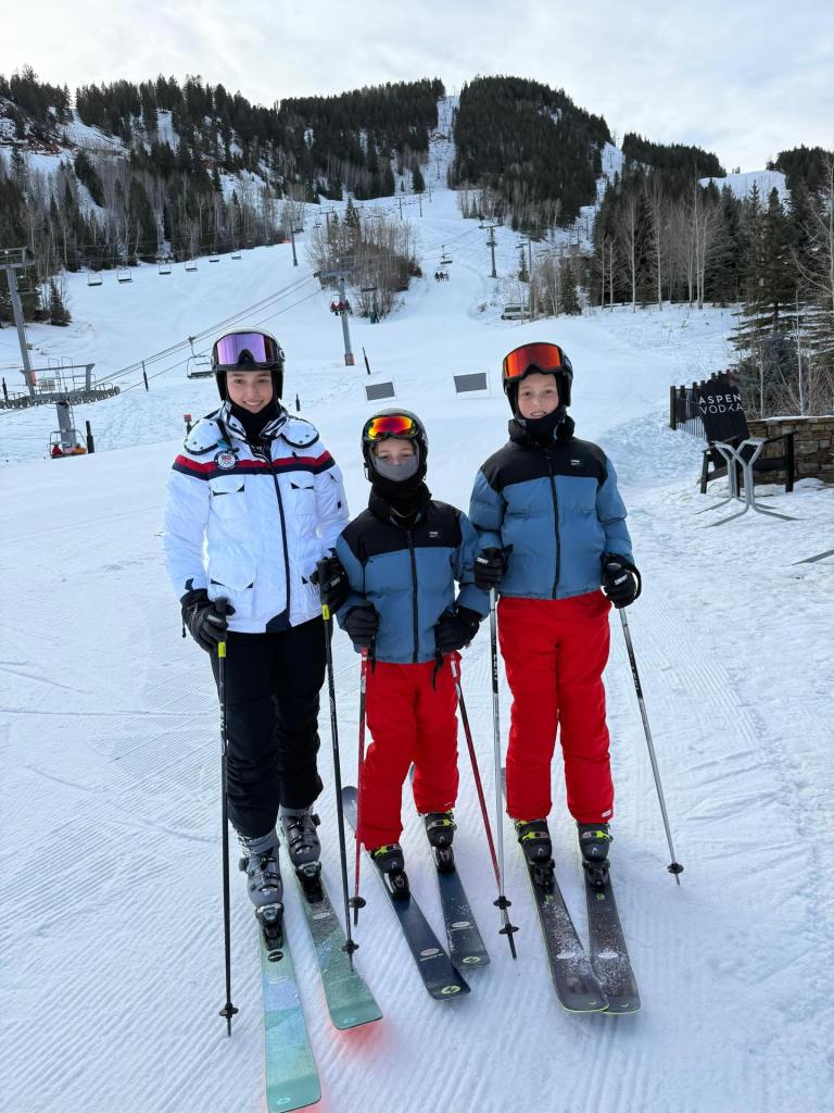 Three children in ski gear stand on a snowy mountain, with ski slopes and trees in the background.