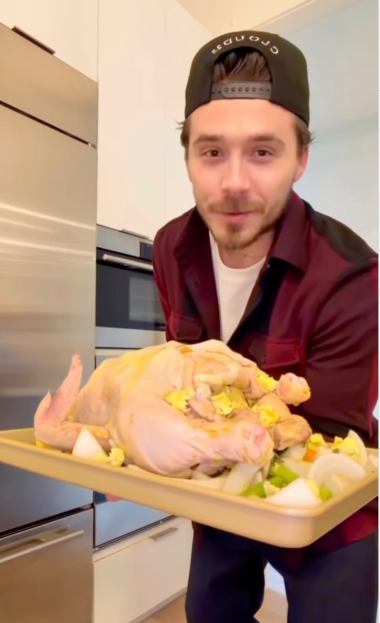 Brooklyn Beckham holding a tray with a raw turkey and vegetables, preparing to cook a roast.