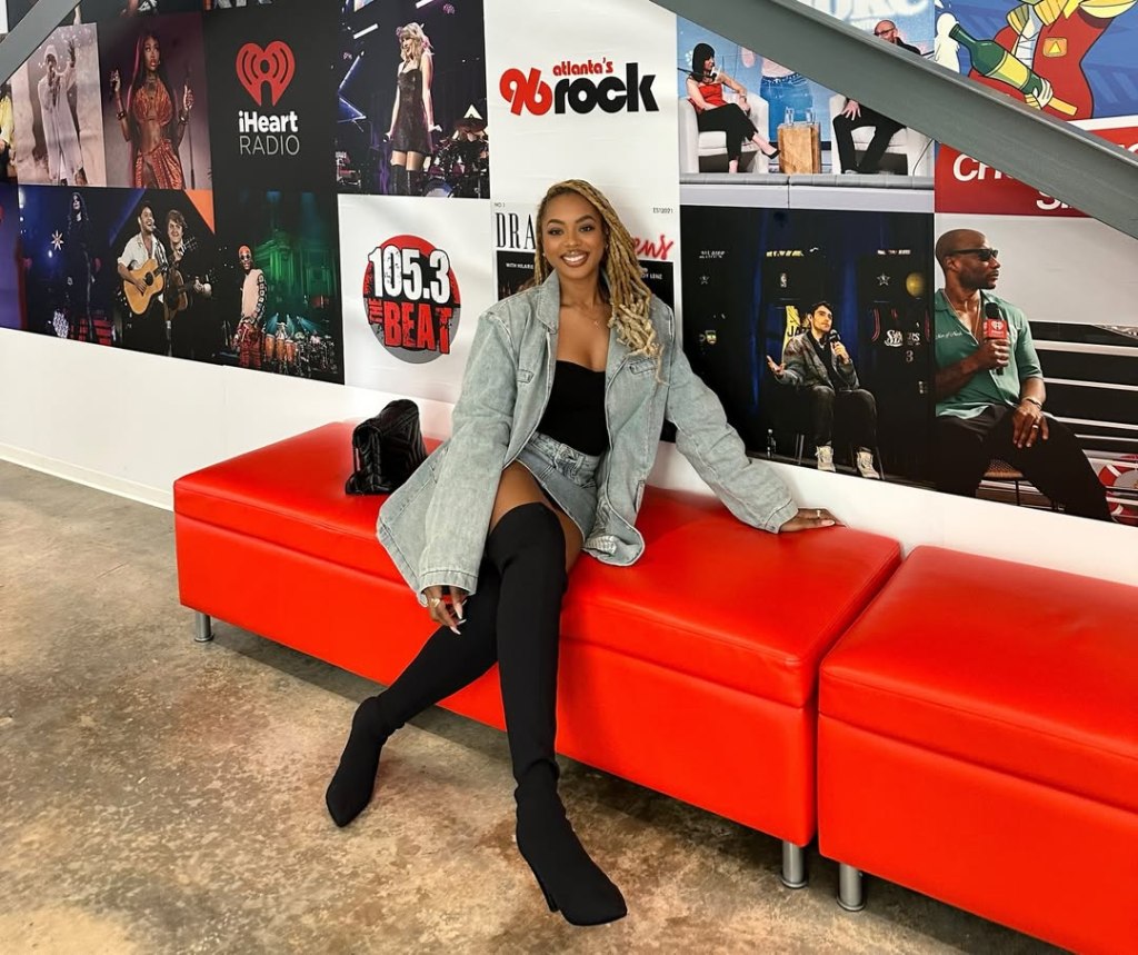 Mickey Lee sitting on a red bench in front of a wall decorated with iHeartRadio and 105.3 The Beat posters.