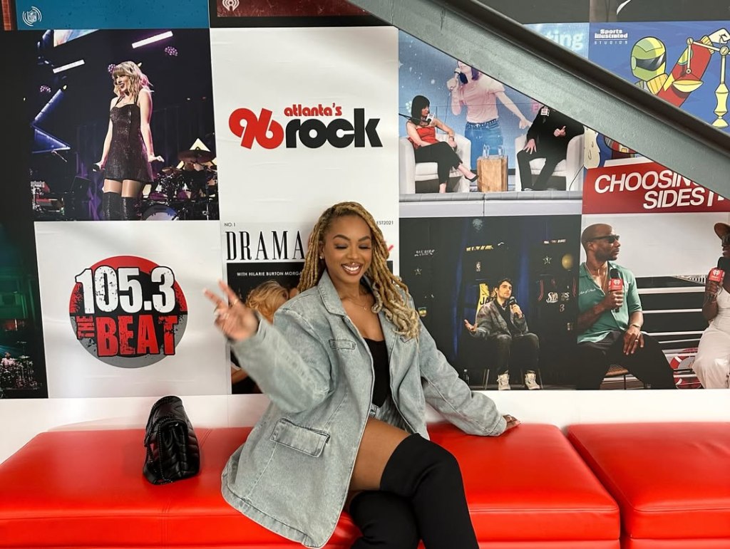Mickey Lee sitting on a red couch, gesturing peace sign, in front of a wall adorned with promotional posters.