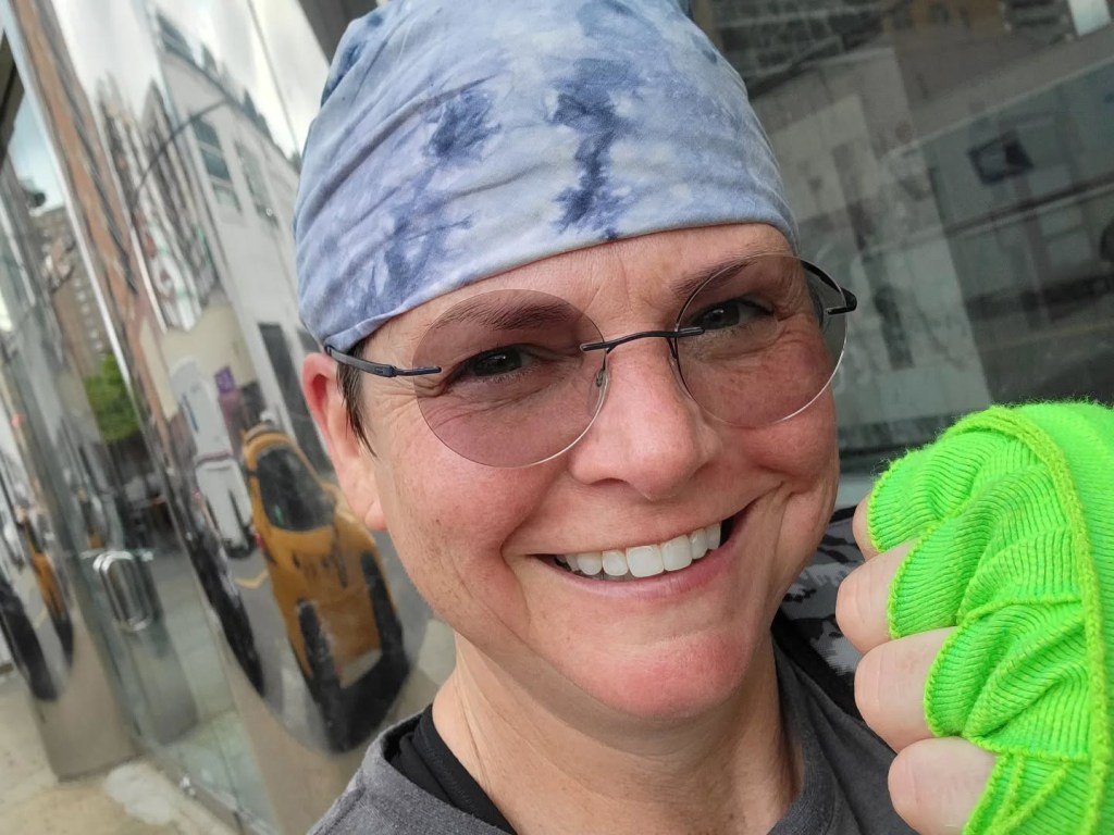 Wenne Alton Davis smiling, wearing a blue tie-dye head wrap, glasses, and a neon green hand wrap, with a distorted reflection of a yellow taxi in the background.