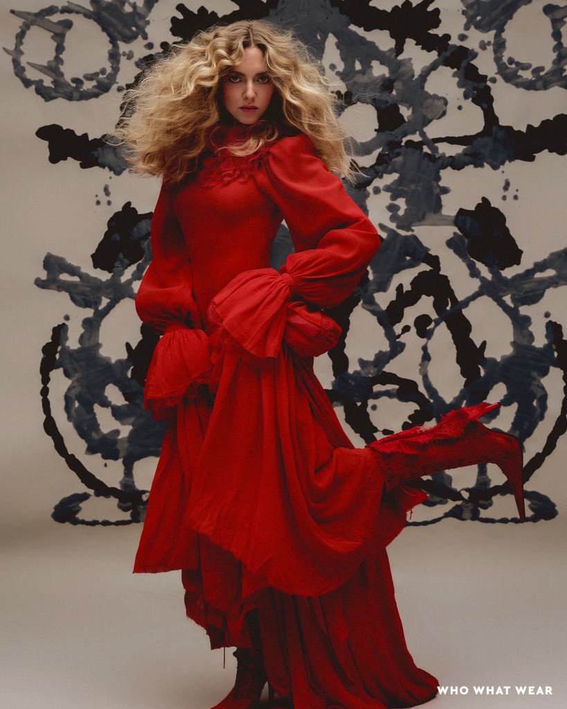 Amanda Seyfried in a red ruffled dress and red boots with a gray and black patterned background.