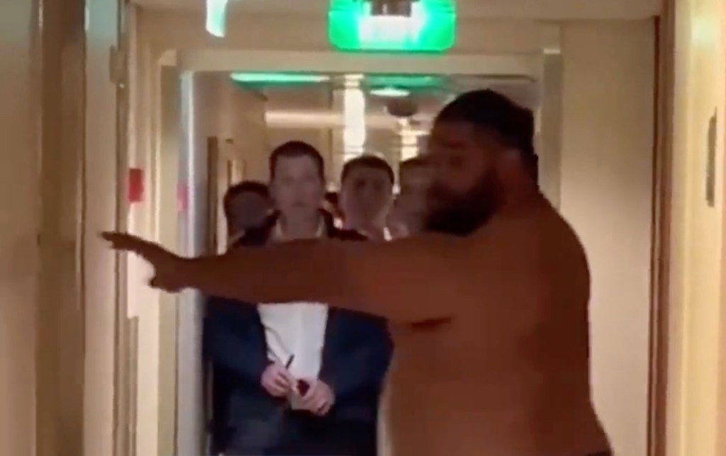 A shirtless man extending his arm, with several men in suits standing behind him in a narrow hallway.