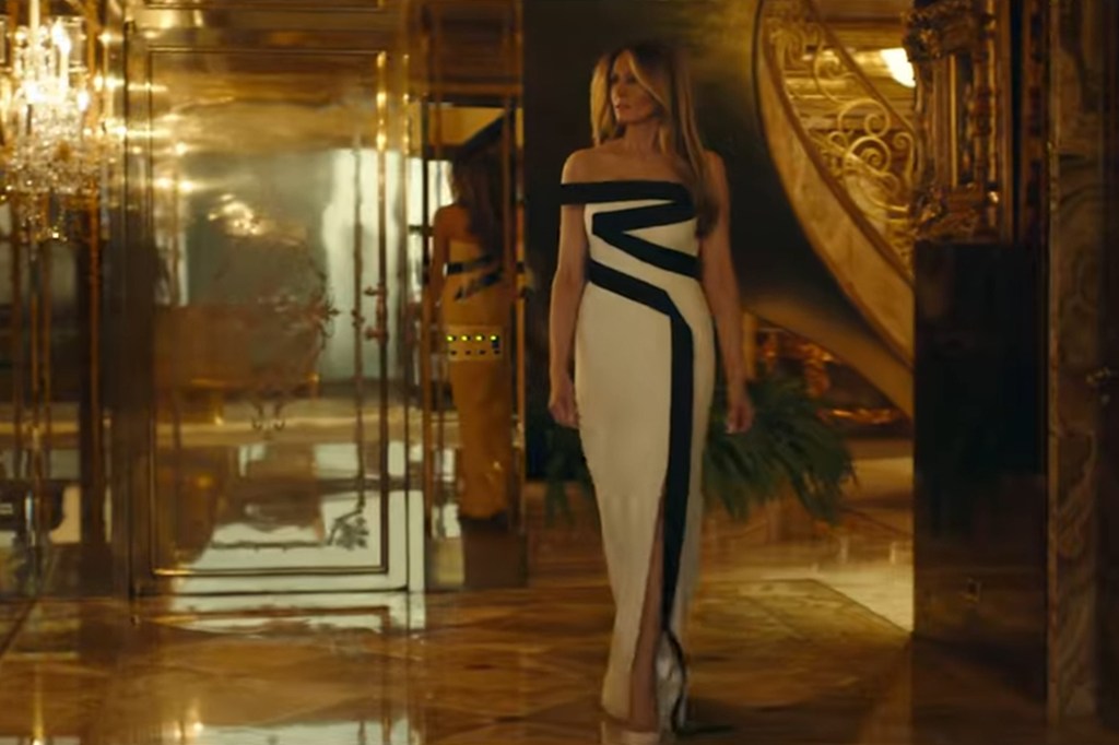 Melania Trump in a white and black gown in a golden room with chandeliers and staircase.