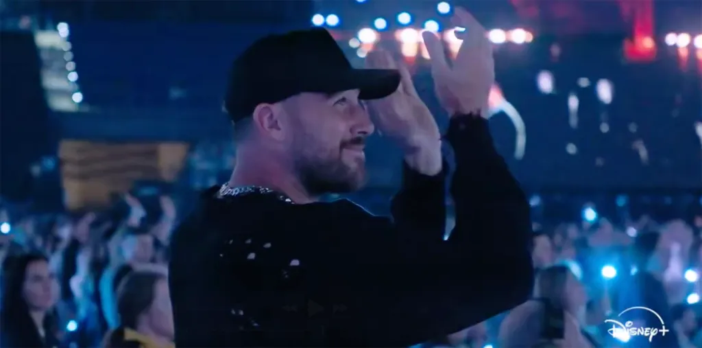 Travis Kelce applauding while wearing a black cap and shirt.