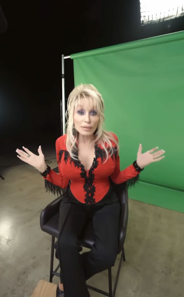 Dolly Parton on set with her hands spread, wearing a red top with black fringes, black pants, and sitting on a stool.