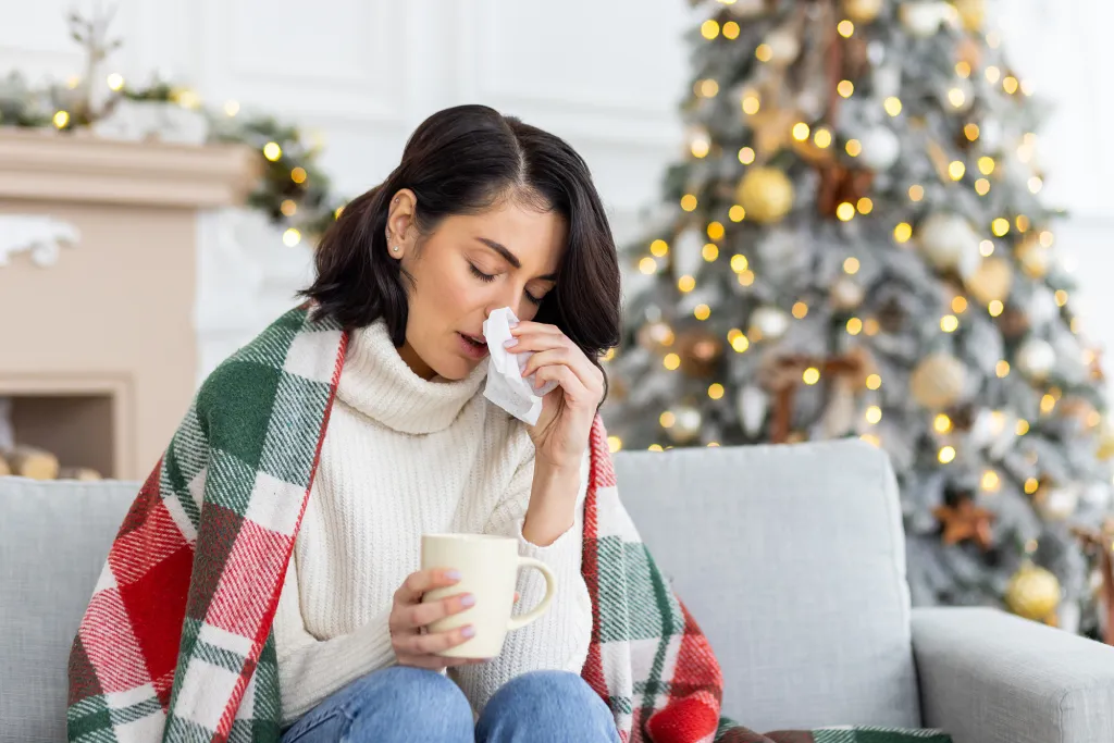 A young woman sits on a sofa, covered with a blanket, holding a mug and wiping her nose with a tissue.