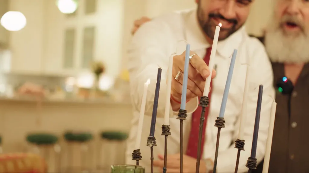 Zohran Mamdani lights a candle in a menorah while Mandy Patinkin looks on.