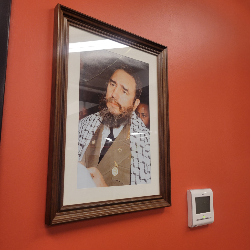 A framed photograph of Fidel Castro in a suit and keffiyeh hanging on a red wall next to a white Fujitsu thermostat.