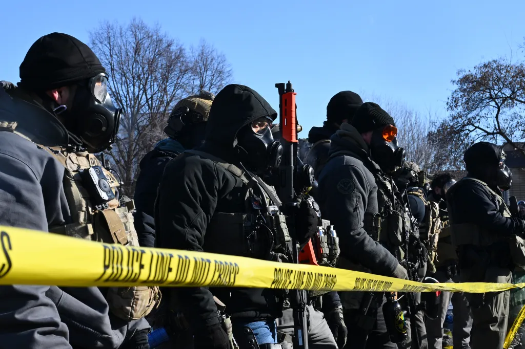 Federal agents in gas masks and tactical gear behind yellow 