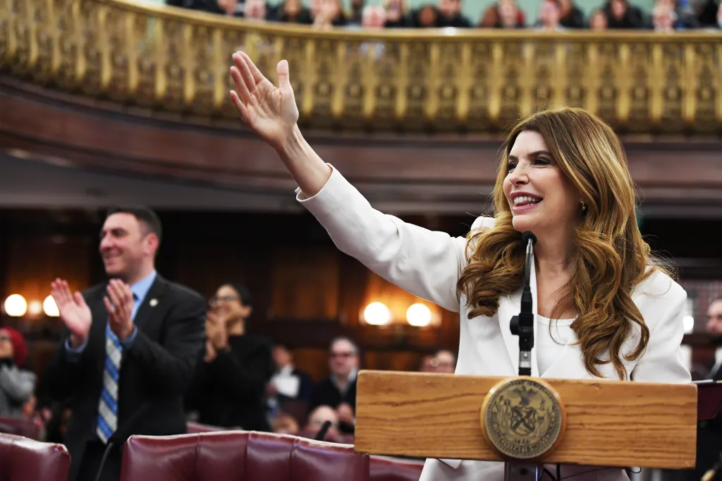 Julie Menin, in a white blazer, waves to an applauding crowd in a legislative chamber.