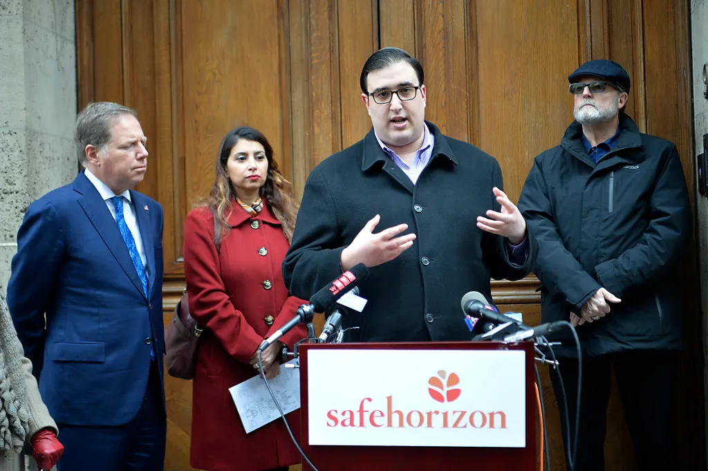 A man wearing glasses and a black coat speaks at a podium with the Safehorizon logo, flanked by two men and one woman.