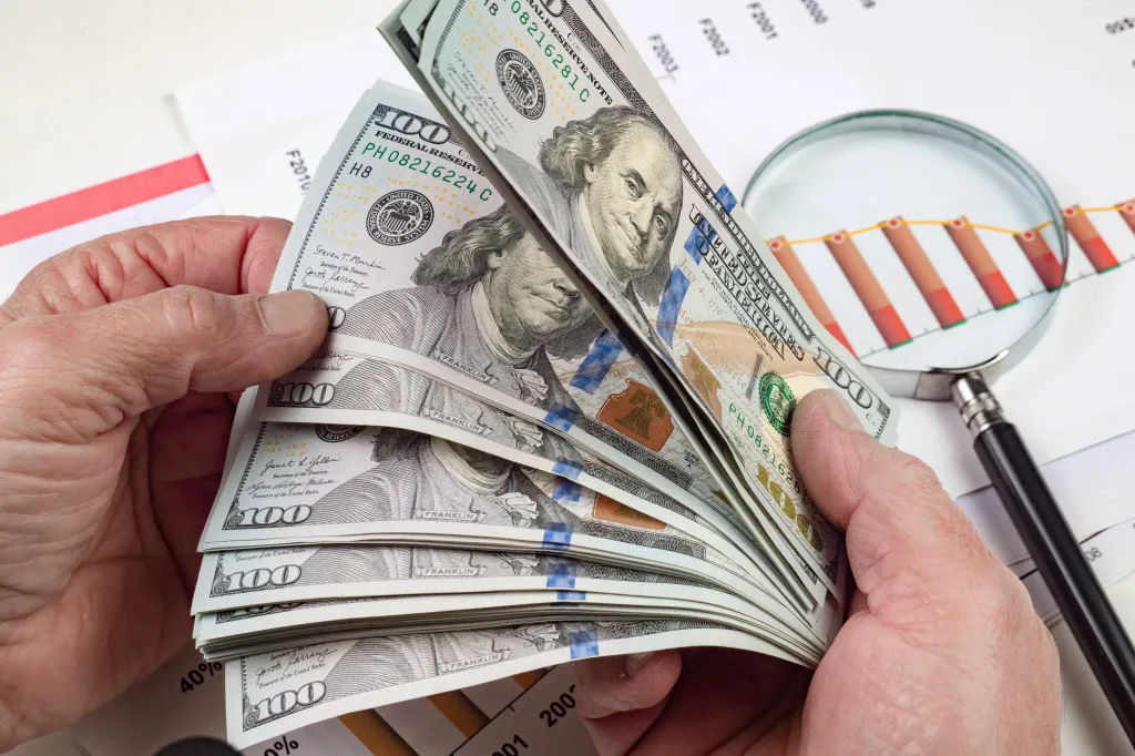 Hands counting a stack of US 100 dollar bills above financial documents and a magnifying glass.