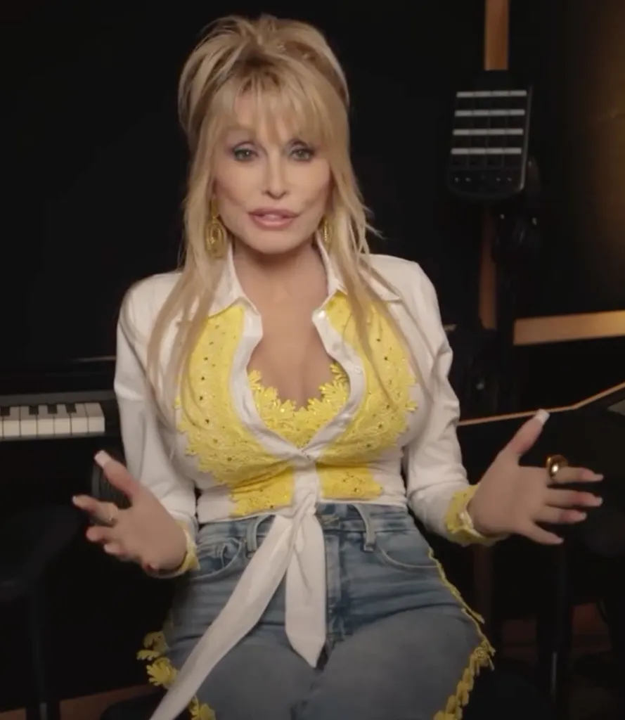 Dolly Parton in a white and yellow shirt and blue jeans, sitting with a keyboard to her left and a bass guitar hanging behind her right.