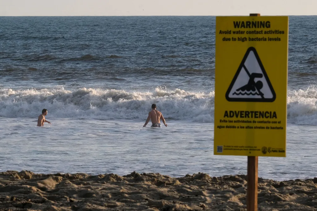 Two men wade in the ocean near a warning sign about high bacteria levels.