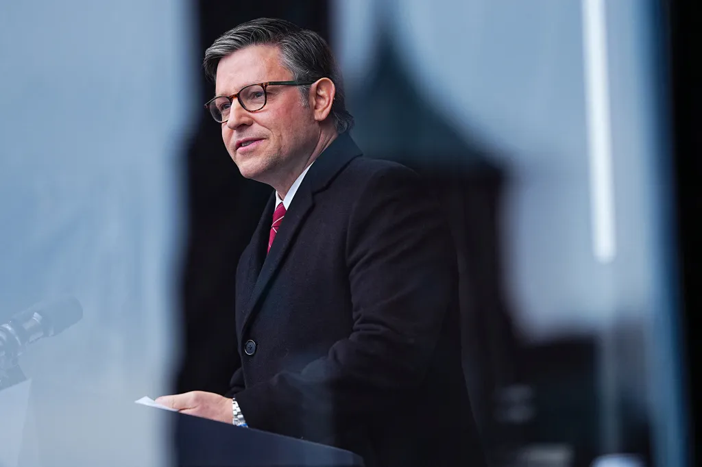 A man with graying hair and glasses wearing a black coat and red tie, speaking at a podium.