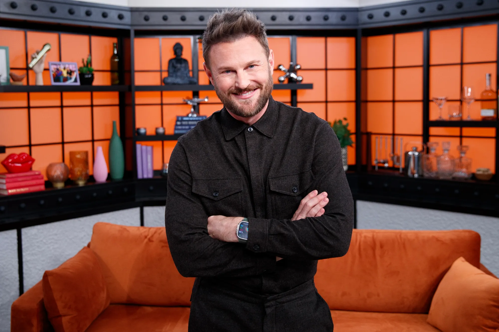 Bobby Berk smiling with arms crossed in front of a modern orange and black shelving unit.