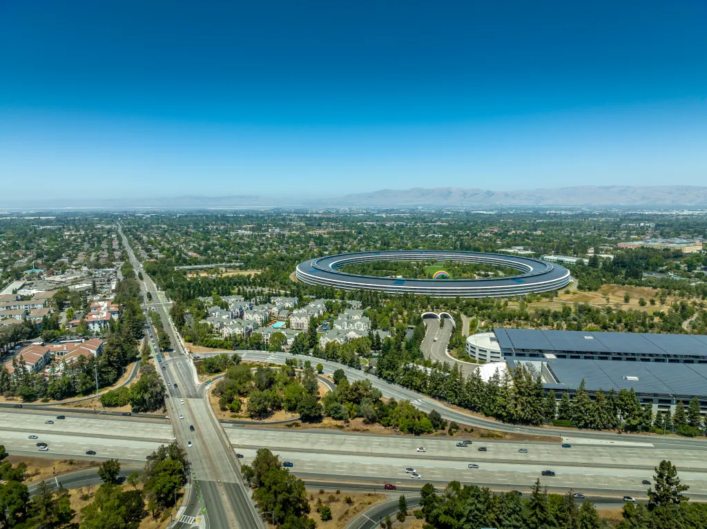 Aerial view of Cupertino, with Interstate 280 running diagonally, past Apple Park's ring-shaped headquarters and a large parking lot.