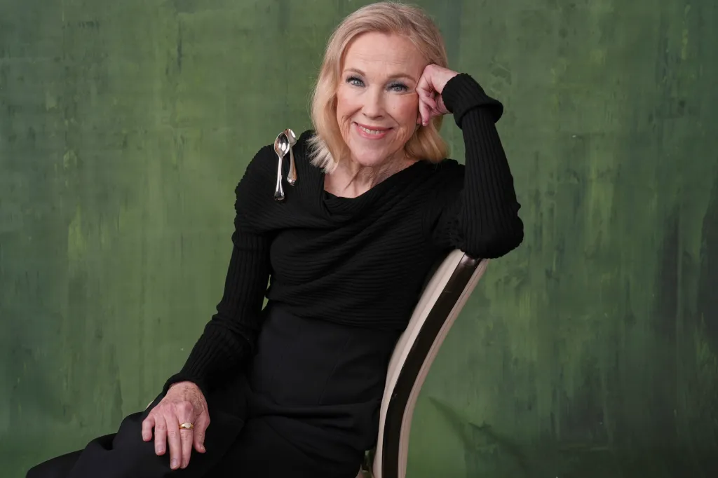 A woman with blonde hair and a black outfit smiles while sitting in a chair against a green background, with two silver spoons pinned to her top.
