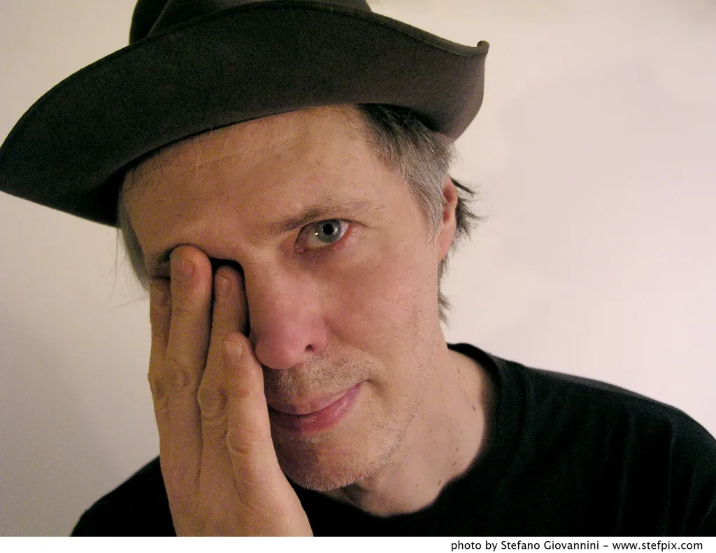 Tom Verlaine wearing a dark hat, with his hand touching his face.