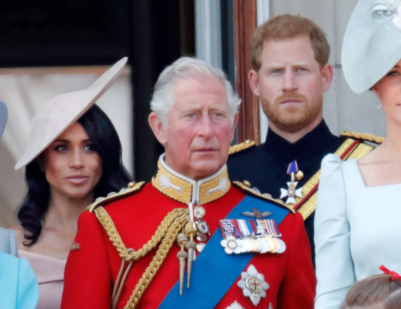 Prince Charles in red military uniform, Meghan Markle in a beige hat, and Prince Harry in dark uniform stand on a balcony.