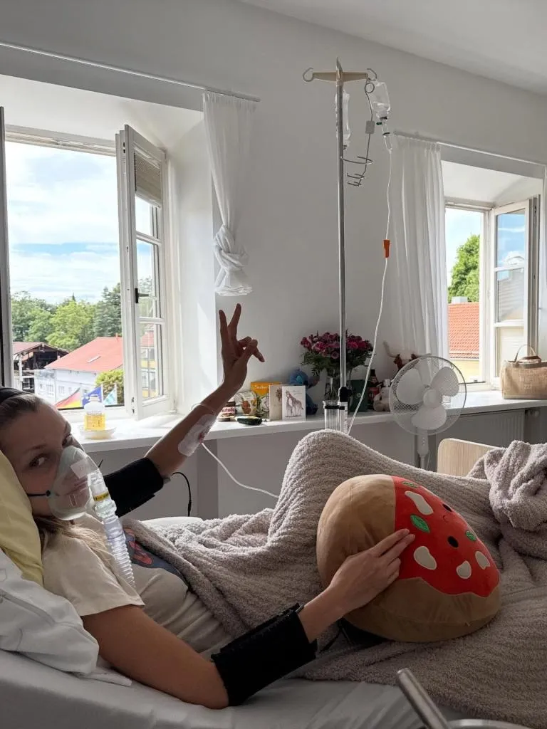 Bella Hadid in a hospital bed with an oxygen mask, making a peace sign, and holding a pizza-shaped pillow.