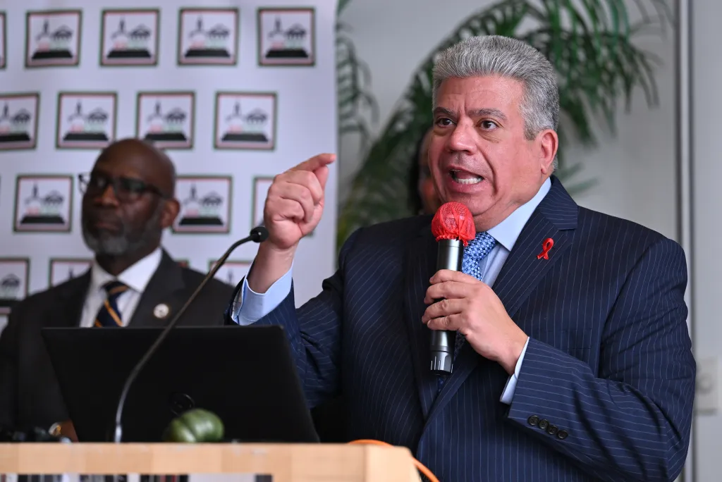 A man in a suit speaking into a microphone, with a man in the background.