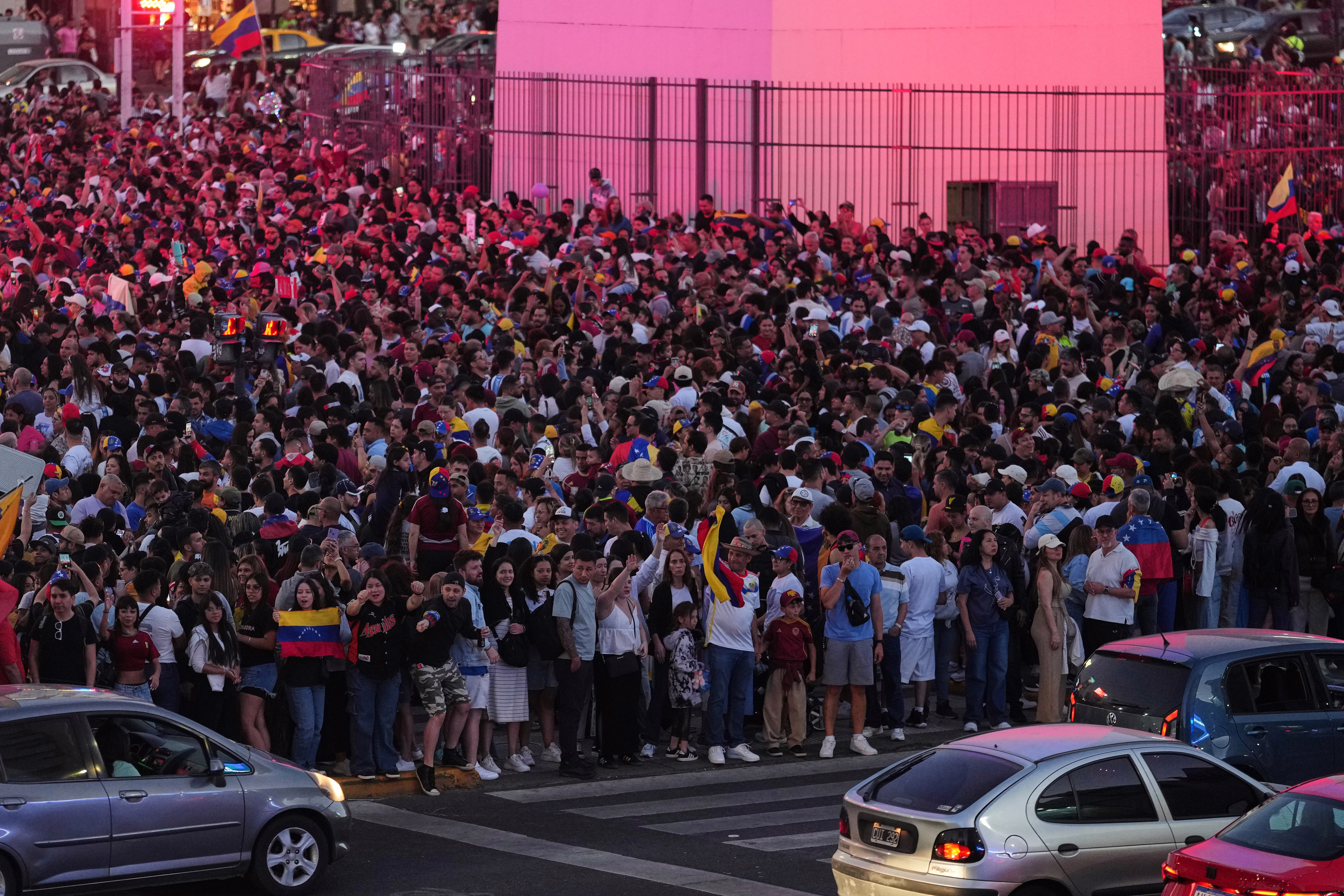 A large crowd gathers in a public square, mini holding Venezuelan flags.