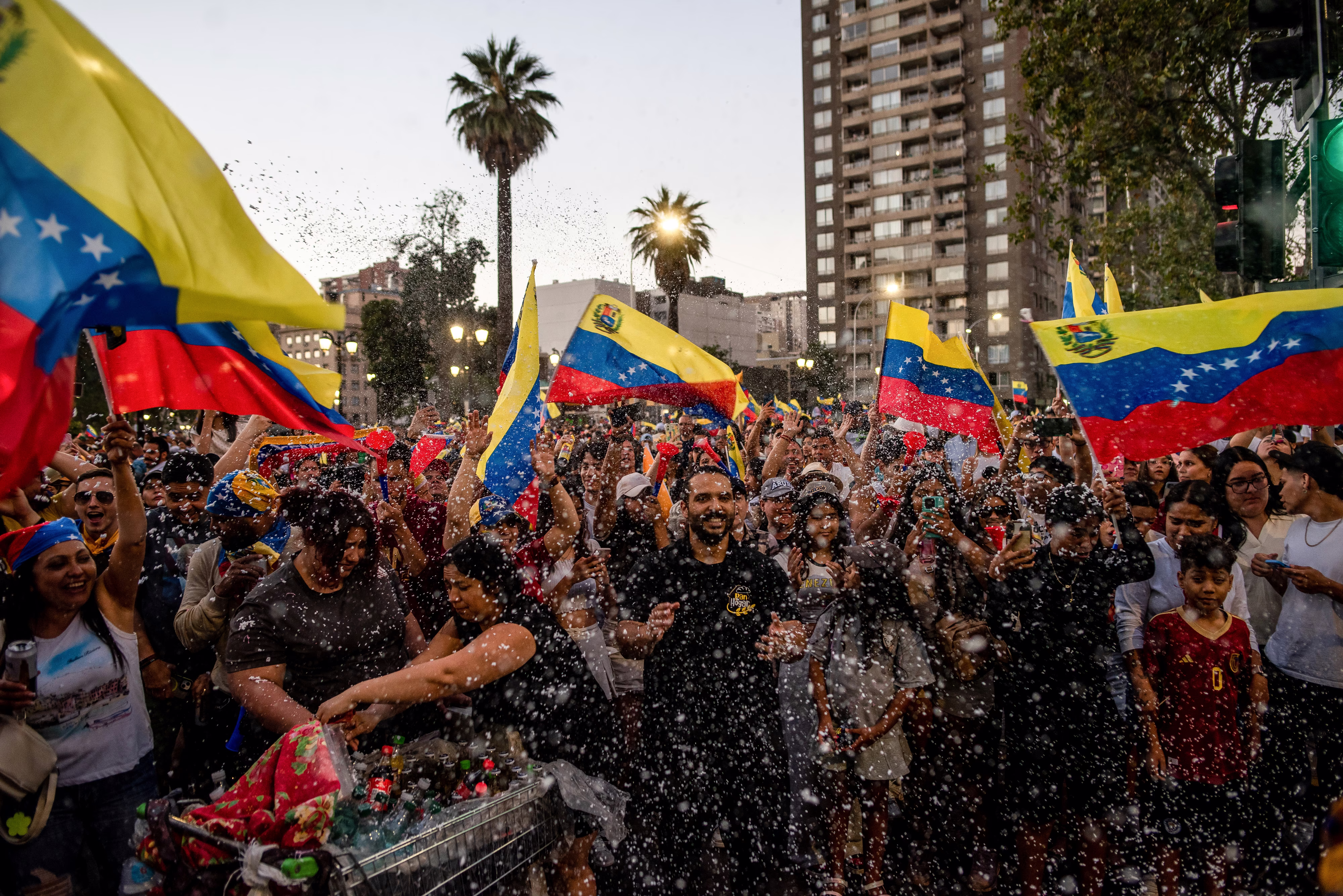 A crowd waving Venezuelan flags celebrates in a public square, near a water fountain.