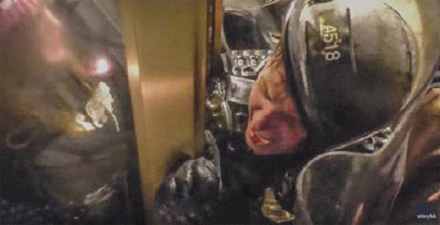 close-up video still of bloodied police officer in riot helmet being crushed in doorway and screaming