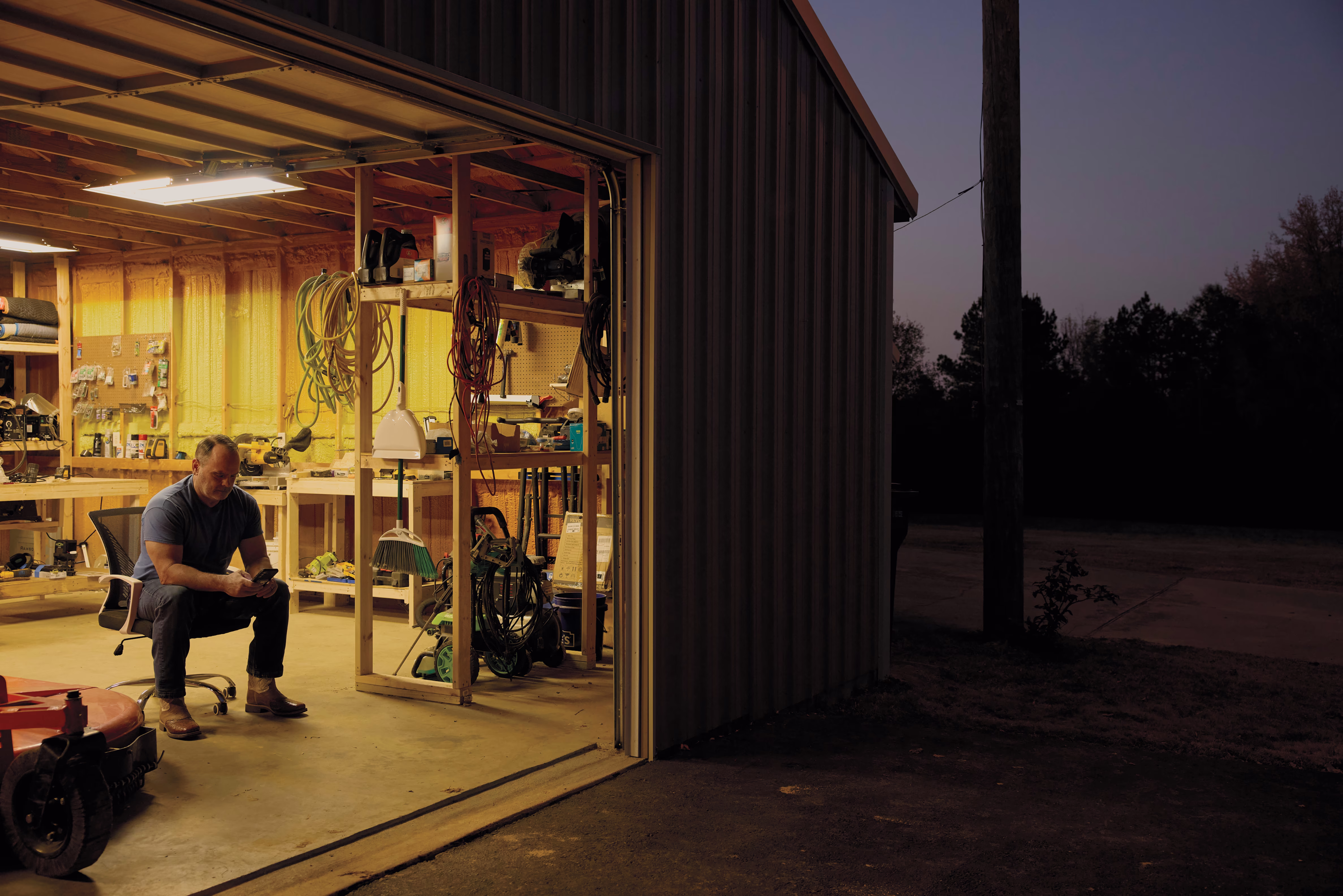 photo of man sitting in doorway of large well lit garage looking at phone at dusk